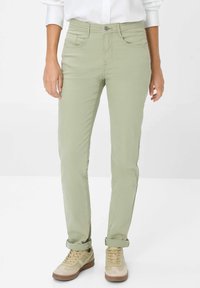 Vorderansicht einer Person in hellgrünen Slim-Fit-Hosen mit hochgekrempelten Säumen und beigen Sneakers, die vor einem weißen Hintergrund steht.