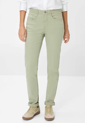 Vorderansicht einer Person in hellgrünen Slim-Fit-Hosen mit hochgekrempelten Säumen und beigen Sneakers, die vor einem weißen Hintergrund steht.