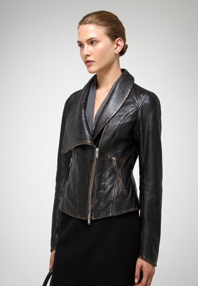 Nainen pukeutuneena istuvaan mustaan nahkatakkiin, jossa on epäsymmetrinen vetoketju ja leveä kaulus, seisoo yksinkertaisen taustan edessä.