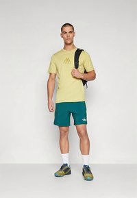 Hele kollane t-särk ja türkiissinised lühikesed püksid logodega. Modell seisab musta seljakotiga. Sinised ja kollased matkajalatsid täiendavad komplekti.