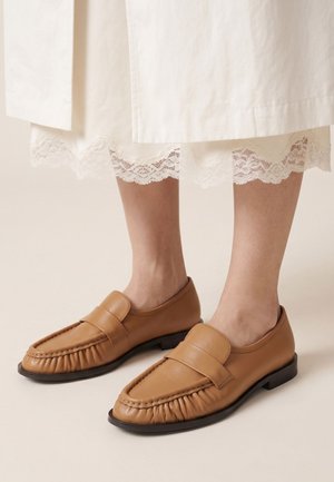 Picioare purtând mocasini din piele cămilă cu detalii strânse, asortați cu o rochie albă cu dantelă la tiv.
