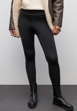 Leggings negros de cintura alta, hechos de un material suave y elástico, combinados con botas negras hasta el tobillo y una chaqueta marrón forrada de borreguito.