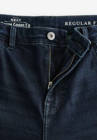 Jeans din denim albastru închis cu fermoar, închidere cu nasture metalic și cusături contrastante. Are o etichetă care indică "Tăietură Normală."