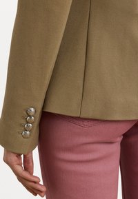 Blazer marron avec texture et boutons argentés aux poignets, associé à un pantalon rose ajusté avec un détail de poche arrière.