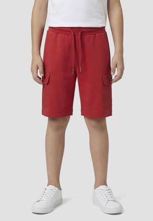 Person trägt rote Cargoshorts mit Kordelzugbund und weißen Sneakers, steht vor einem schlichten hellen Hintergrund.