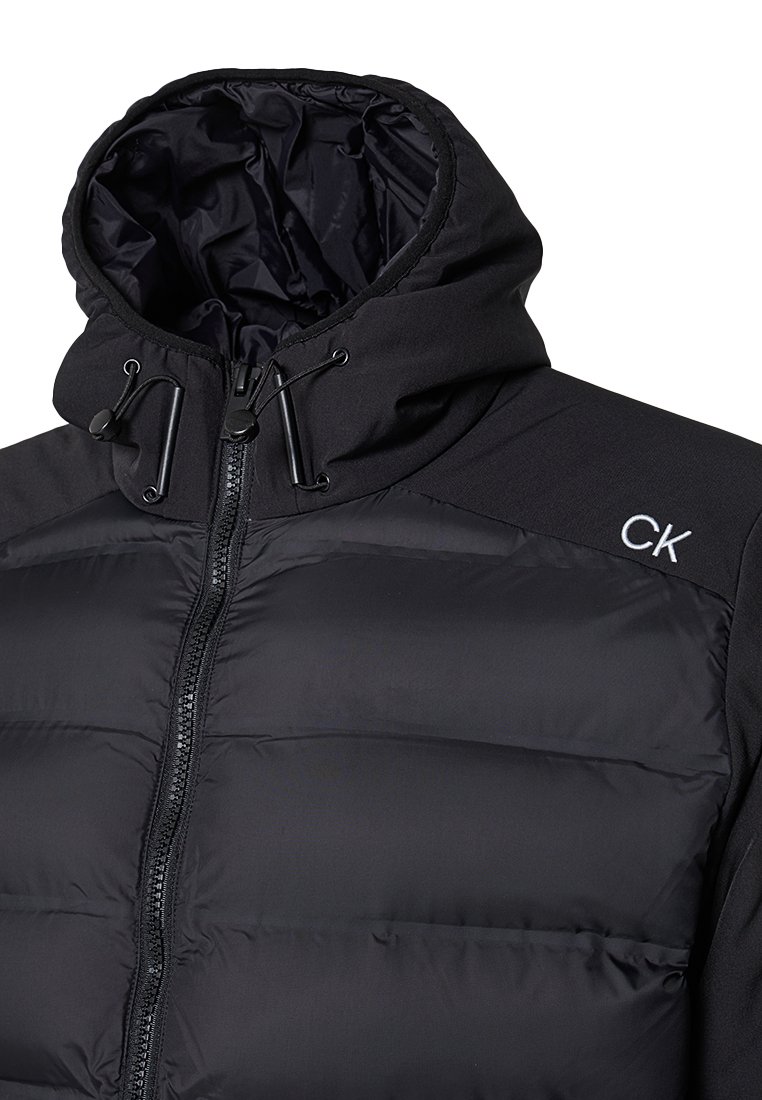 Czarna, ocieplana kurtka z kapturem, z przodu na zamek błyskawiczny, z regulowanymi sznurkami i logo CK na ramieniu. Gładka, wodoodporna tekstura.