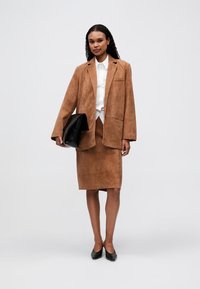 Vrouw in een bruine suède blazer en rok met een wit shirt, met een grote zwarte clutch in de hand en zwarte hakken aan, staande op een effen achtergrond.