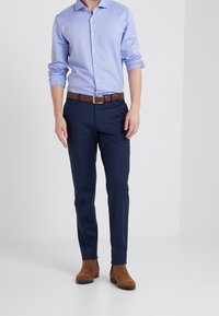 Man in een lichtblauwe button-up shirt met opgerolde mouwen, marineblauwe broek, bruine leren riem en bruine suède enkellaarzen die op een witte vloer staat.
