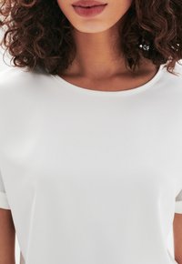 Top blanco de manga corta con cuello redondo, textura suave y diseño minimalista. La tela parece ligera y ligeramente reflectante.