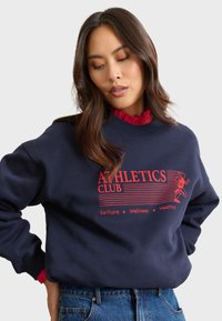 Marineblaues Sweatshirt mit rotem "ATHLETICS CLUB"-Druck, ausgestattet mit Streifen und Wellness-Slogans. Strukturiertes Gewebe, gerippte Bündchen und Rundhalsausschnitt.