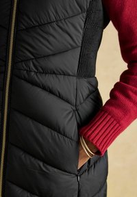 Czarna kamizelka puchowa z pikowanym wzorem i złotym zamkiem, połączona z czerwonym swetrem z prążkowanymi mankietami. Ubranie ma detal w postaci bocznej kieszeni.