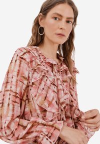 Průsvitná růžová halenka s abstraktními hnědými vzory, volánkovým výstřihem a manžetami, dlouhými rukávy a šňůrkami vpředu. Viditelný detail texturované látky.