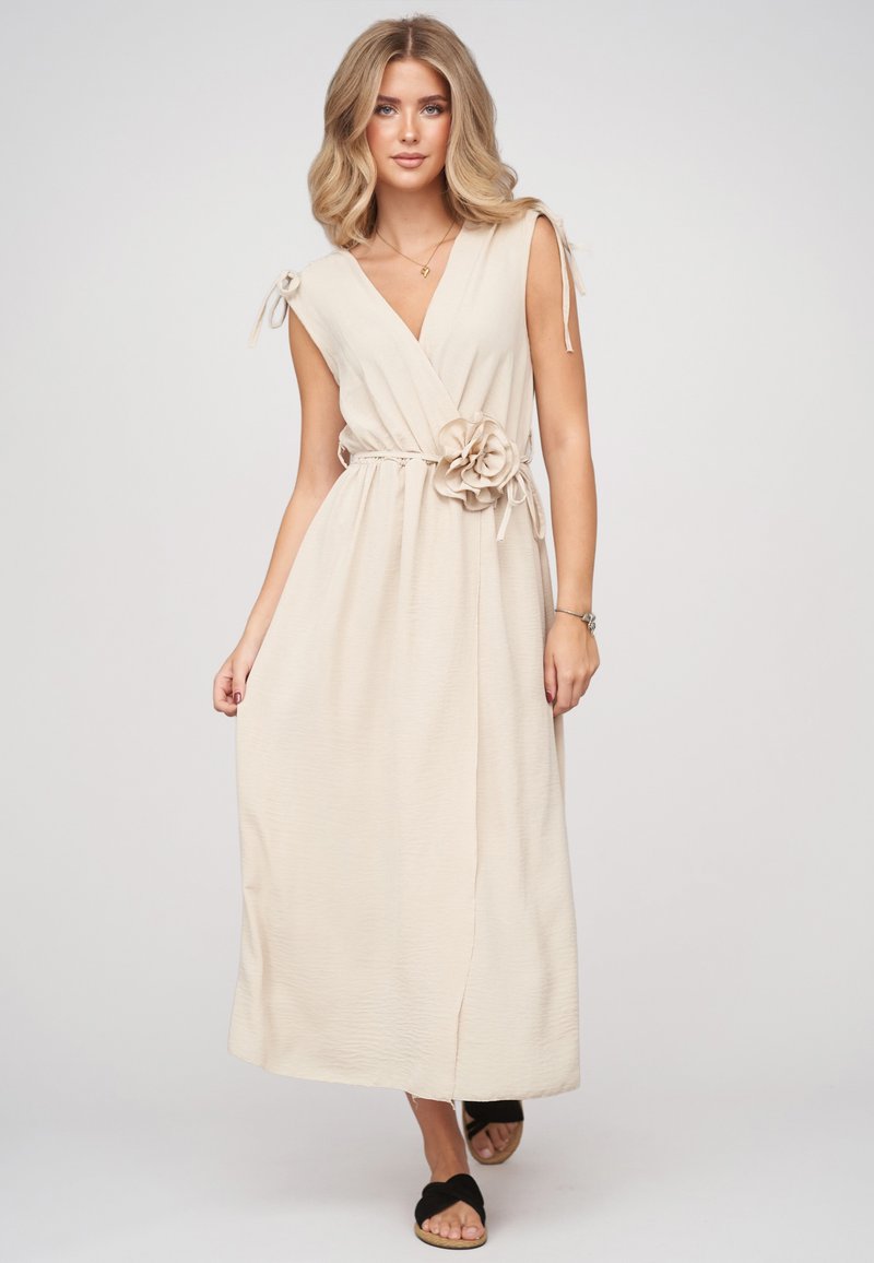 Femme aux cheveux blonds porte une robe portefeuille beige sans manches avec une fleur en tissu à la taille et des sandales noires faciles à enfiler.