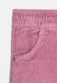 Tissu en velours côtelé rose tendre avec des nervures verticales, doté d'une taille élastique et d'une poche latérale avec un détail de bouton-pression en métal.