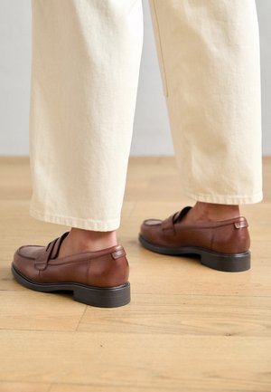 Brązowe skórzane buty wsuwane z niskim czarnym obcasem, o teksturowanym wykończeniu i minimalistycznym designie, połączone z szerokimi spodniami w kolorze kremowym.