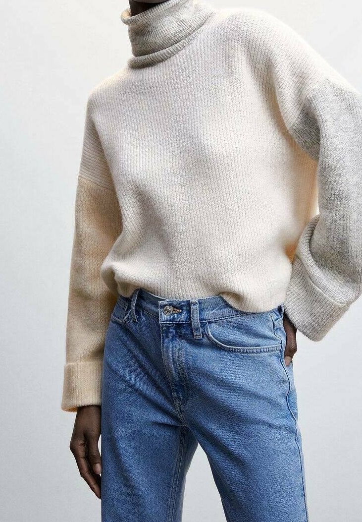 Personne portant un pull à col roulé côtelé couleur crème avec des manches bicolores rentré dans un jean taille haute bleu clair.