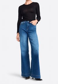 Schwarzes, langärmeliges, geripptes Oberteil, kombiniert mit hochgeschnittenen, weit geschnittenen blauen Jeans mit leichter Ausbleichung und umgeschlagenem Saum. Schwarze, hohe Schuhe.