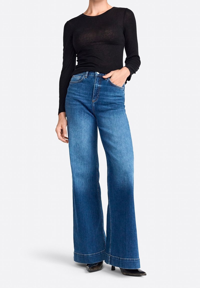 Schwarzes, langärmeliges, geripptes Oberteil, kombiniert mit hochgeschnittenen, weit geschnittenen blauen Jeans mit leichter Ausbleichung und umgeschlagenem Saum. Schwarze, hohe Schuhe.