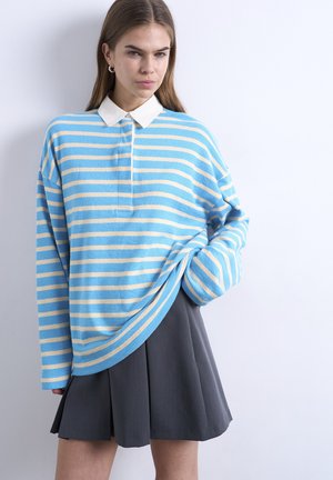 Jeune femme portant un haut à manches longues rayé bleu et beige avec un col blanc et une jupe plissée gris foncé, debout contre un mur blanc.