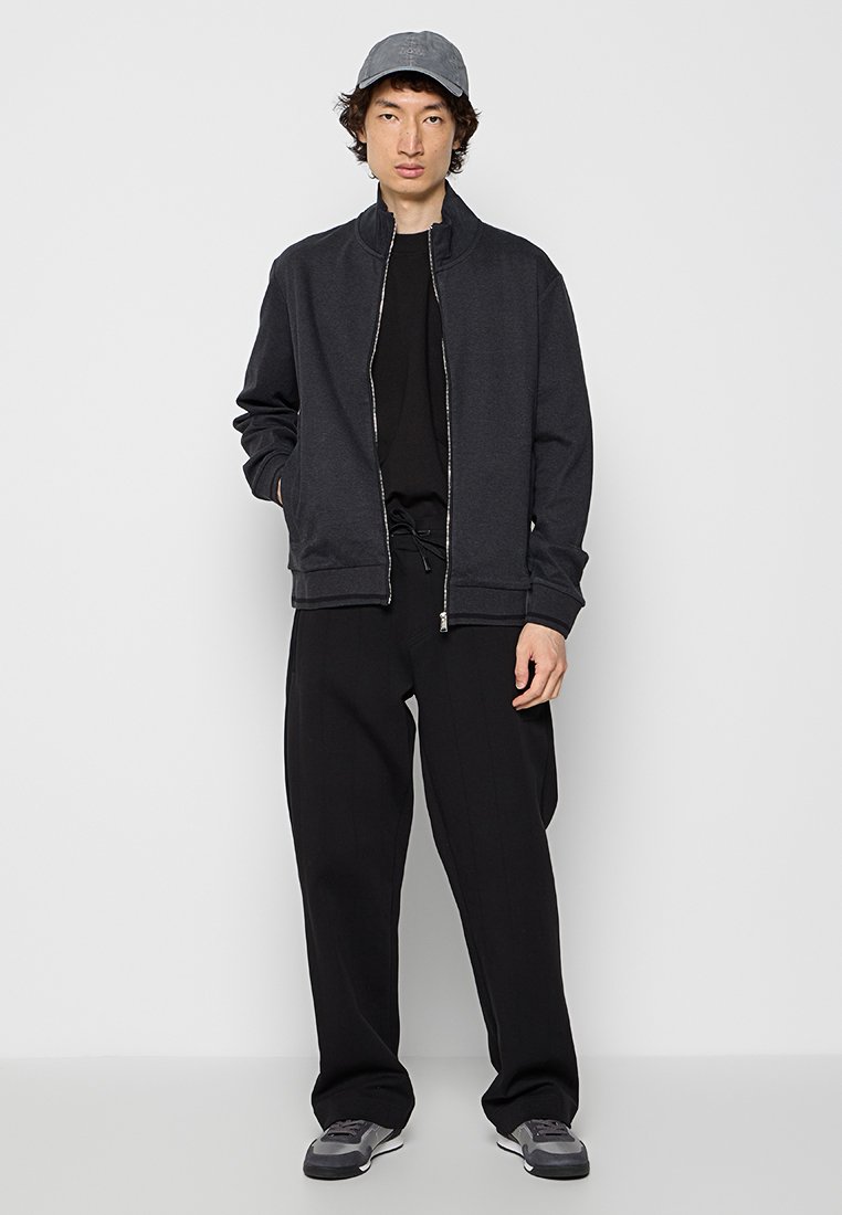 Jonge man met een grijze pet, zwart zip-up jack, zwart shirt, brede zwarte broek en grijze sneakers staat tegen een simpele witte achtergrond.