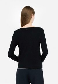 Mujer con cabello castaño liso, vistiendo una camiseta negra de manga larga con un logo sutil en el cuello y un ribete plateado en el dobladillo, de espaldas.