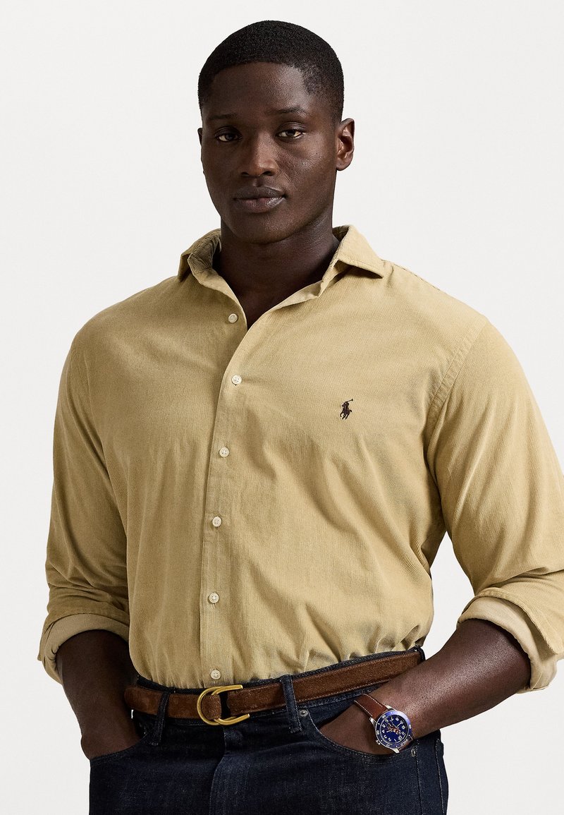 Homem vestindo uma camisa de corduroy bege com mangas arregaçadas, jeans escuros, cinto castanho e um relógio de pulso com mostrador azul e bracelete de couro castanho.