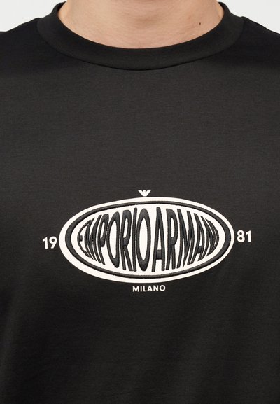 Μαύρο T-shirt με λευκό οβάλ λογότυπο Emporio Armani, κείμενο "1981" στις πλευρές και τη λέξη "Milano" τυπωμένη κάτω από το λογότυπο.