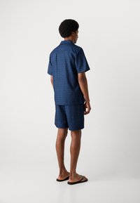 Kurze Ärmel, Hemd mit Kragen und passende Shorts in Dunkelblau, mit einem subtilen geometrischen Muster und einer lässigen Passform. Flache Sandalen runden den Look ab.