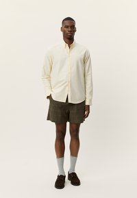 Ανοιχτό κίτρινο πουκάμισο με κουμπιά, χακί σορτς με σχέδια, γκρι κάλτσες και καφέ παπούτσια, στέκεται μπροστά σε ουδέτερο φόντο.