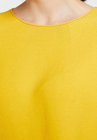 Gelbes Strickoberteil mit rundem Halsausschnitt, strukturiertem Stoff und sichtbaren Nähten. Das Material scheint leicht und atmungsaktiv zu sein.