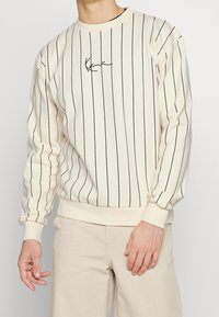 Sudadera crema con rayas verticales negras, cuello redondo, puños acanalados y logo bordado en el pecho. Se lleva con pantalones beige claros.
