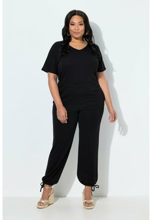 Žena s dlouhými tmavými vlasy, oblečená v černém krátkém topu a černých kalhotách s vázáním u kotníků, stojící na bílém pozadí v zlatých sandálech na klínu.