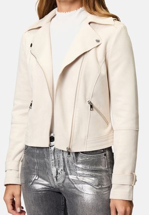 Kobieta ubrana w jasnobeżową zamszową kurtkę biker z srebrnymi zamkami błyskawicznymi i metalicznie szare spodnie dżinsowe o kroju do pasa.