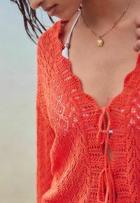 Robe de plage en crochet orange avec un décolleté à volants et un lien, arborant un motif de dentelle délicat. Portée par-dessus un haut de bikini.
