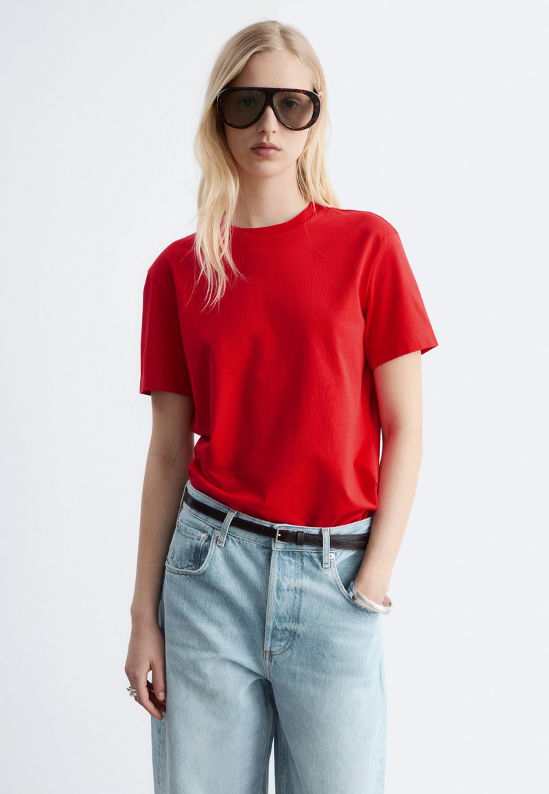 Femme blonde portant de grandes lunettes de soleil noires, une chemise rouge à manches courtes rentrée dans un jean bleu clair taille haute avec ceinture noire, une main dans la poche.