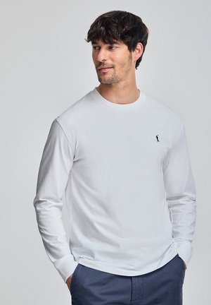 Witte langslavenshirt van katoen, met een ronde hals en een klein zwart logo op de borst. Soepele textuur, eenvoudig ontwerp.