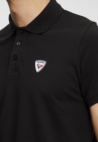 Černé polo tričko s texturovanou tkaninou, s límcem a třemi knoflíky. Na levé straně hrudi se nachází nášivka s logem Rossignol v červené, bílé a modré barvě.