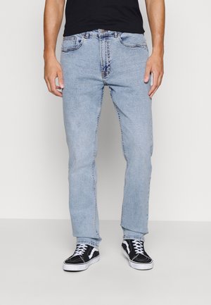 Homme portant un jean bleu clair et des baskets noires avec des lacets blancs, debout devant un fond clair uni.