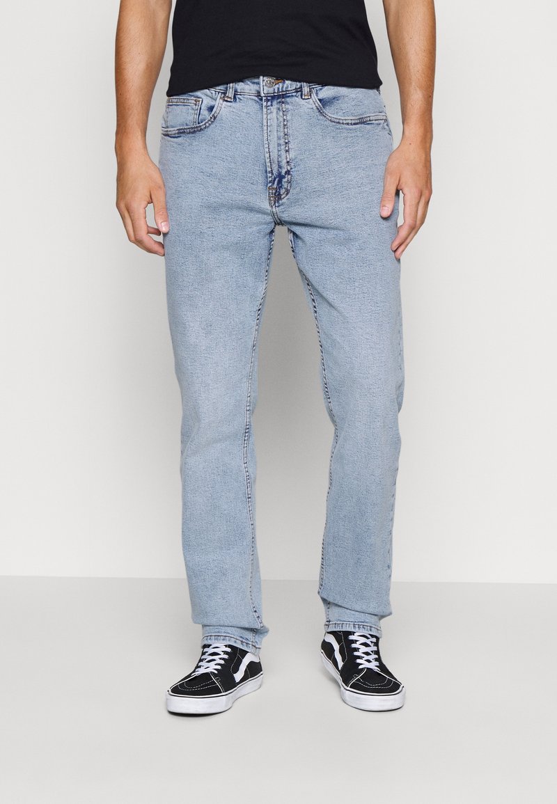Homme portant un jean bleu clair et des baskets noires avec des lacets blancs, debout devant un fond clair uni.