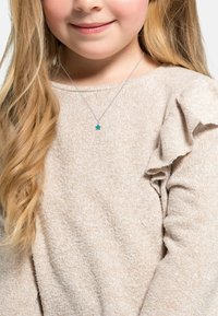 Beige, texturerad tröja med volangdetalj på axlarna. Silverkedja med ett litet, turkoshjärta hänge. Långt blont hår.