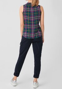 Donna in piedi, che indossa un top senza maniche a quadri verde, viola e bianco, pantaloni cargo blu navy arrotolati e sneaker bianche con dettagli argentati.