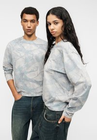 Ανοιχτόγριζα φούτερ με αφηρημένα μπλε σχέδια, συνδυασμένα με σκούρα τζιν. Μαλακό ύφασμα, μακριά μανίκια, άνετη εφαρμογή, μινιμαλιστικός σχεδιασμός.