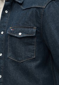 Μπροστινή τσέπη μπουφάν από σκούρο μπλε τζιν με καπάκι, πορτοκαλί ραφές, ασημένιο κουμπί με σούστε και μικρή υφασμάτινη ετικέτα σε κόκκινο-άσπρο-μπλε χρώμα.