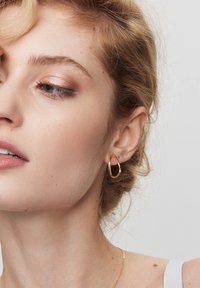 Złote kolczyki w formie dużych kół z wydłużonym kształtem kropli, o gładkiej fakturze i wypolerowanym wykończeniu. Noszone na uchu modelki.