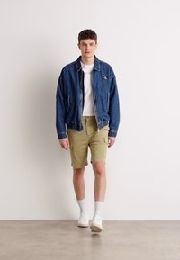 Denim jacket with logo, white t-shirt, olive cargo shorts, white sneakers, and ankle socks. Model stands against a plain background.