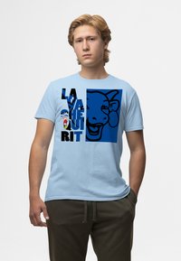 Světle modré tričko s grafikou kravské tváře v modré a černé barvě, s textem "LA VACHE QUI RIT" a logem na hrudi.