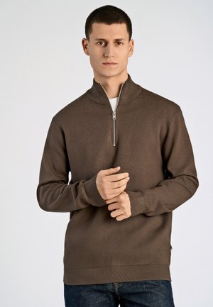 Brązowy sweter zapinany na zamek z wysokim kołnierzem, wykonany z gładkiego materiału. Posiada długie rękawy i dopasowaną talię. Nosi się na białej koszuli.