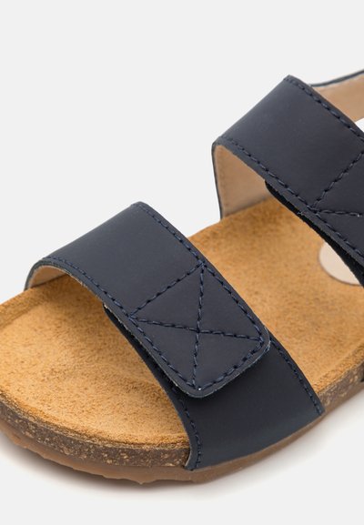 Temno modri sandal z dvema širokima naramnicama, šiviljskim detajli in plitvim podplatom iz plute. Teksturni gumijasti podplat za oprijem in podporo.