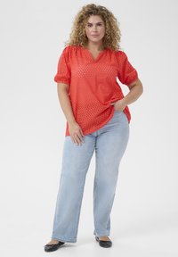 Blouse rouge semi-transparente avec un motif à pois, manches courtes bouffantes et col en V, associée à un jean droit bleu clair et des ballerines noires.