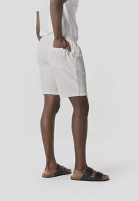Persona con pantalones cortos blancos con textura, bolsillo trasero y sandalias negras con hebilla, de pie con una mano en el bolsillo sobre un fondo liso.